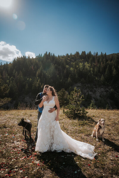 A groom hugs his bride from behind as they stand on a grassy hillside with a mountain behind them, and they're gazing off to the left. Their two dogs sit on either side of them.