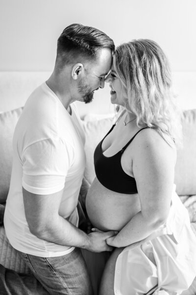 A black and white image of a pregnant couple sitting closely together. They are forehead to forehead, smiling warmly, as the man gently holds the woman’s belly.