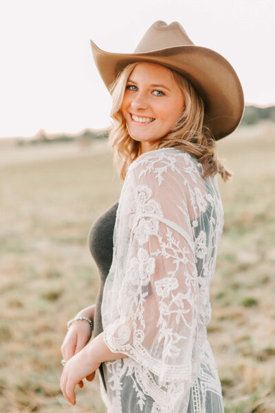 A high school senior in a cowboy hat, dress, and lace duster smiles and looks over her shoulder