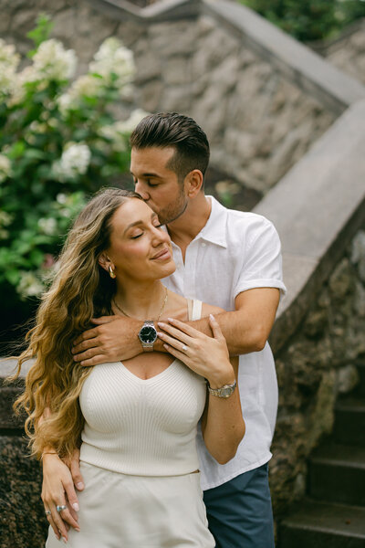 A man gently embraces a woman from behind, kissing her head. They stand on stone steps, surrounded by greenery and flowers. The woman has long curly hair and smiles with closed eyes, wearing a cream outfit, while the man wears a white shirt.