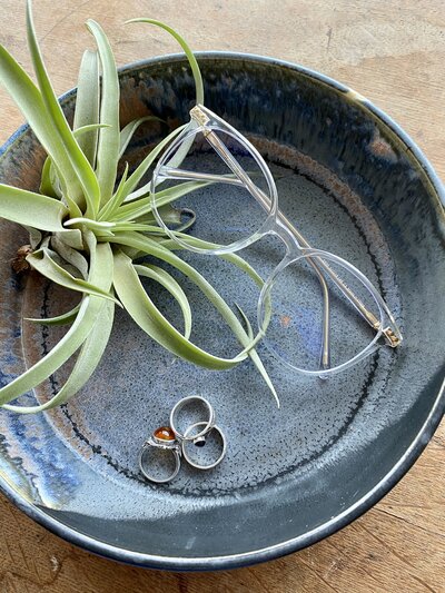 A pair of blue light glasses sits in a pottery bowl alongside an air plant and three rings.