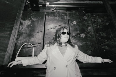 Black and white photo of a women in a suit jacket with sunglasses, arms splayed on a long table behind her, looking off to the side