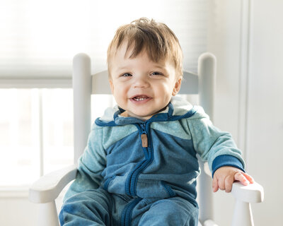 12 month old boy with short dark hair, sitting a light gray rocking chair, smiling off to the side of the camera, wearing a blue zip up one piece outfit