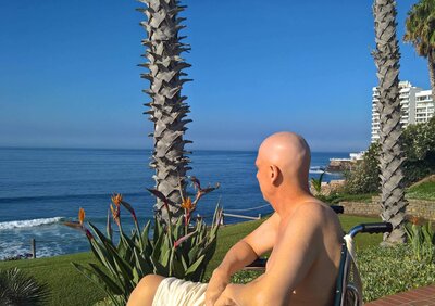 Eric Leseberg sitting in a wheelchair enjoys a seaside view, surrounded by palm trees and modern buildings, perhaps imagining himself within the pages of a Phillipa Leseberg book.