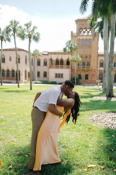 A couple sharing a romantic kiss on a grassy lawn with a large, Mediterranean-style historic estate in the background. The man, wearing a white shirt and dark trousers, dips the woman, who is wearing a long yellow dress, while palm trees frame the architecture.