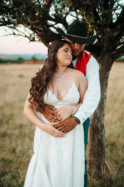 BRIDE  WITH CLOSED EYES LEANING ON GROOM WHILE HE WRAPS HER  FROM BEHIND  IN OPEN FIELD WITH A TREE in san antonio, texas.