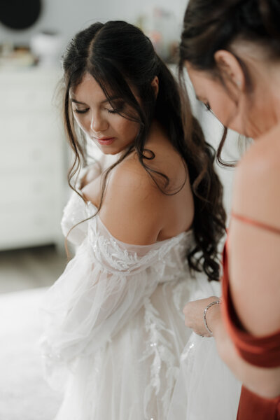 In a softly lit room, captured by a New England wedding photographer, a bride with long, dark hair and a white dress glances over her shoulder. A woman in red gracefully assists with the gown, embodying the essence of fine art weddings and portraits in New England.