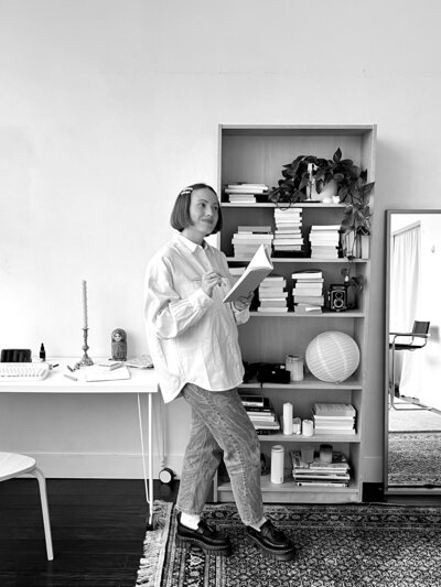 A greyscale photo of margarita standing in front of a bookshelf with a notebook open, slightly smiling as she catches a spark of creativity.