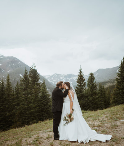 bride and groom are hiking with mountain views as the backdrop. It is summer, and they off roaded in the mountains after saying their vows at an alpine lake with family and friends. The bride is holding her dress as she stands next to the groom in a black suit.