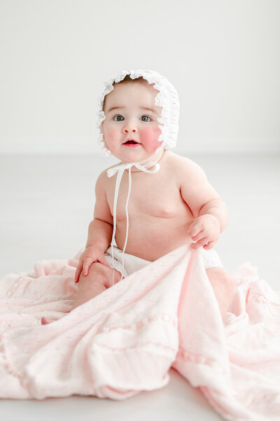 a baby girl sits on a pink blanket wearing a white bonnet