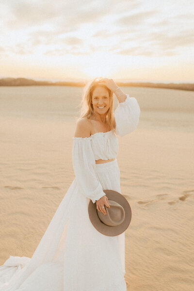 Blonde woman  looking right at the camera, she is wearing a long white skirt with a an off the shoulder matching top. She is on the beach at sunset.