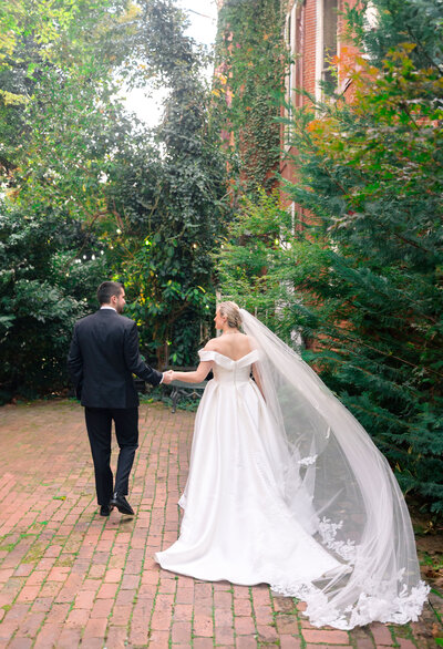 A couple walks hand in hand down a brick path surrounded by lush greenery. The bride wears an elegant white gown with a long veil, and the groom is in a black suit. The scene is peaceful and romantic, set in an outdoor garden.