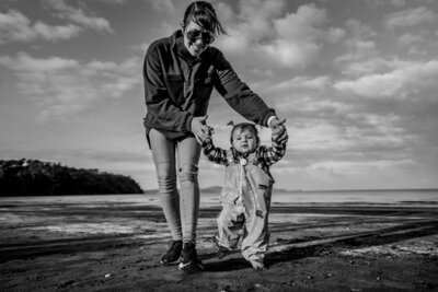 Black and white photo of Zanthe Vorsatz holding her daughter’s hands as she takes baby steps on the beach during a natural family photoshoot in Auckland.