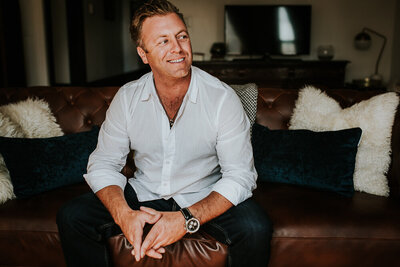 Handsome blonde man wearing a white button-down shirt and dark jeans sits on a brown leather couch with legs spread slightly and forearms resting on knees and hands clasped