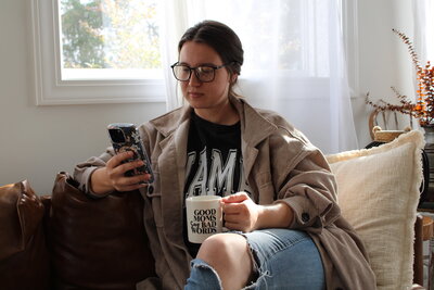 The Comma Mama Co working on her phone wearing a black Mama tee and holding a "good moms say bad words" coffee mug.