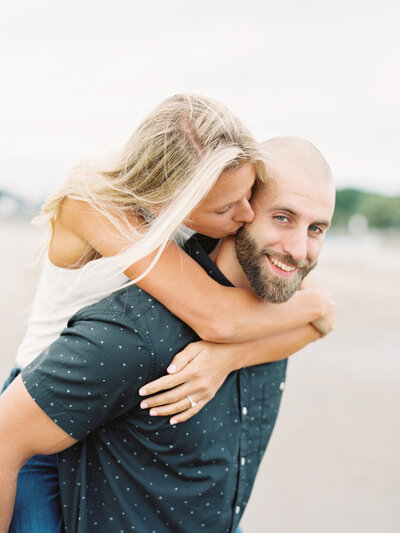 Engagement photo of a bald man carrying his fiance with blond hair on his back while she kisses him on the cheek in Portsmouth, New Hampshire