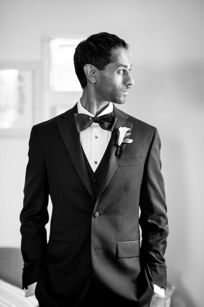 A man in a black tuxedo with a bow tie stands confidently, looking to his right. He has a boutonniere on his lapel. The black-and-white photo captures him in a well-lit room with soft focus in the background.