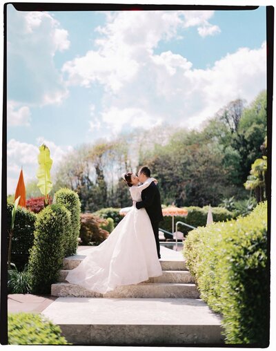 A bride and groom share a kiss on a stone balcony in villa del Balbianello overlooking Lake Como, captured by an Italy wedding photographer. The brides gown extends into a long, elegant train, effortlessly blending with the leafy branches and potted plants that frame this picturesque scene.