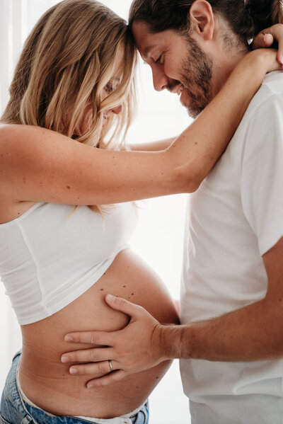Mom and dad embrace while looking down at pregnant belly.