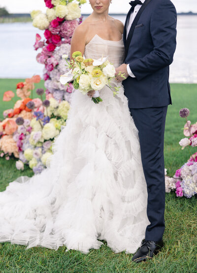 A bride in a flowing white dress holds a bouquet of yellow and white flowers. She stands with the groom in a black suit and bow tie. They are outdoors near a lake, surrounded by colorful floral arrangements.