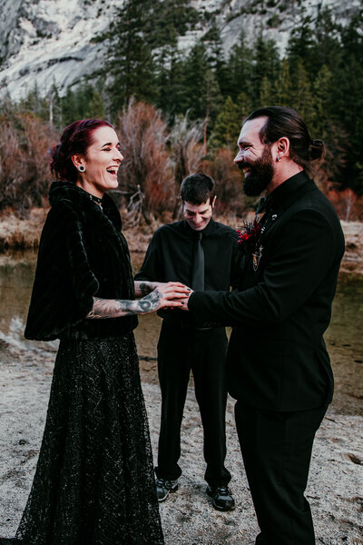 Bride and groom laughing during their elopement ceremony at Yosemite National Park, with the officiant standing behind them near a serene water setting.