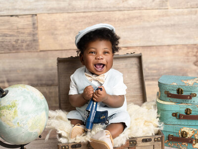 A joyful toddler in a gray romper sits on a white sofa surrounded by cookie-themed decor. The scene includes blue balloons and a plush cookie monster.