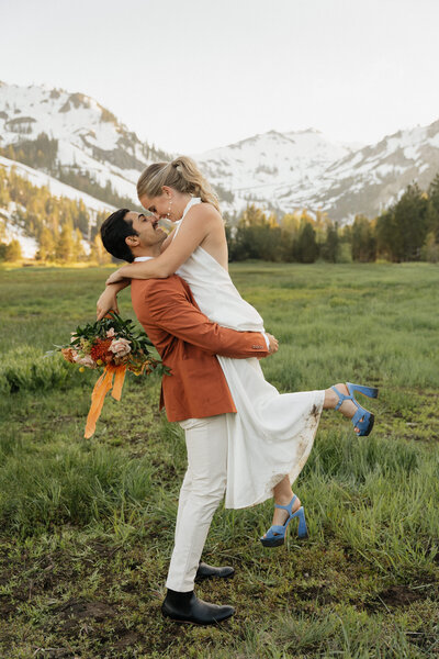 groom picking bride up off the ground while standing in a field with trees and mountains in the background