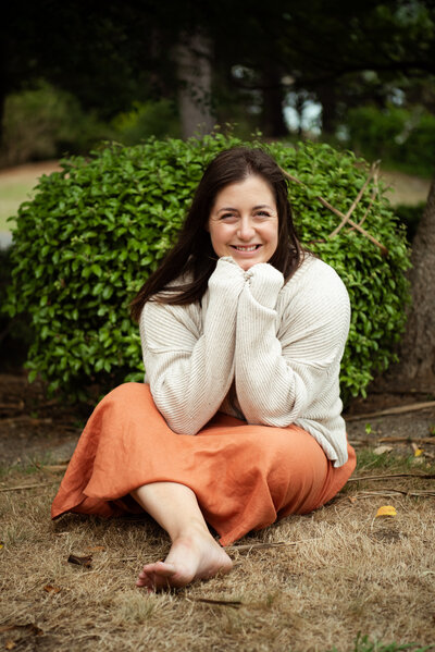 Chantal sitting in front of a lush green bush, barefoot on dry grass, smiling softly while bundled in a cozy cream sweater and orange skirt - Family Autism Support Coach at Chantal Hewitt Coaching