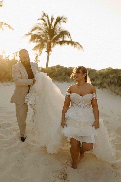 Groom holding the train of bride’s dress as they walk along the beach at sunset, with palm trees in the background.