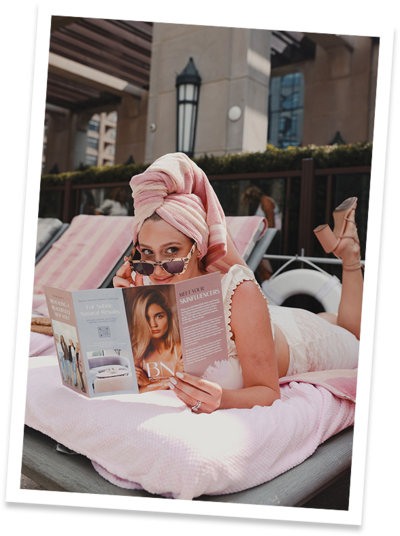 A woman in a pink bathing suit and a pink towel on her head as she relaxes by the pool with a fashion magazine in hand during a social media photoshoot directed by Mello Social.