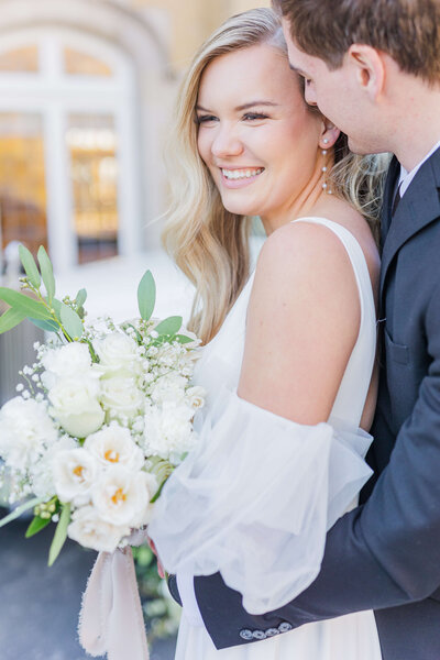 A bride and groom huge each other as she smiles at the camera and he has his arms around her and has his nose by her check. She is holding her bouquet full of white roses.