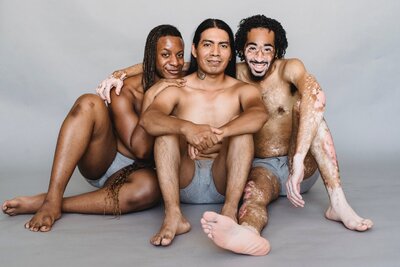 Three masculine presenting people of color wearing gray boxers sit together on the ground, arms wrapped around each other.