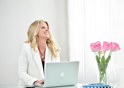 woman in white blazer looking out the window with big smile on laptop