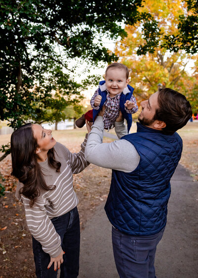 Lifestyle photo of parents holding their laughing son in the air at an Essex County, New Jersey park in the fall.