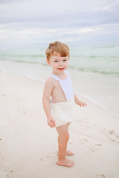 A joyful young boy runs barefoot along the sandy beach, his face beaming with happiness. Dressed in a classic white and yellow romper, he plays near the gentle ocean waves, capturing the carefree essence of childhood in a light and airy family portrait by a Birmingham family photographer."