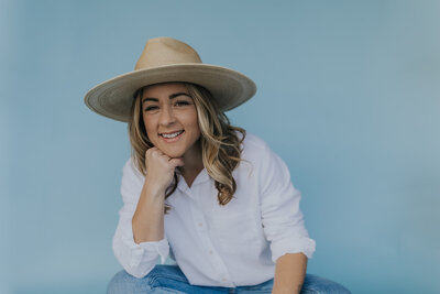 Owner of Peaches and Poppies Floral wearing a cute hat, white cotton shirt, smiling at the camera in front of a blue background
