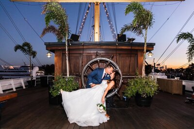 A romantic dip kiss aboard The Moshulu in Philadelphia – Under the twilight sky, the groom sweeps the bride into a dramatic dip kiss in front of the ship’s wooden captain’s wheel. String lights illuminate the rigging above them, and the bride’s flowing wedding dress cascades behind her as she clutches a bouquet of white roses. The historic ship, docked along the Philadelphia waterfront, provides a uniquely romantic backdrop