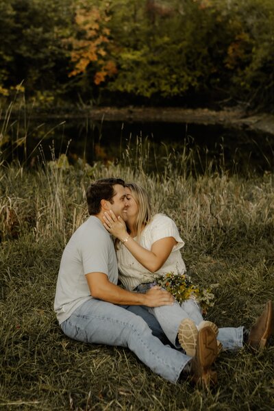 An engaged couple sits on the ground together, holding one another close.
