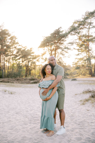 Een stralend stel geniet van een liefdevol moment op het strand, met de zwangere vrouw in een elegante witte jurk en haar partner beschermend achter haar.