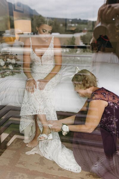 A bride in a lace wedding gown looks down lovingly at her mom who is putting on her bridal shoes at the St. Julien in Boulder, CO. Photo by Haley Hawn.