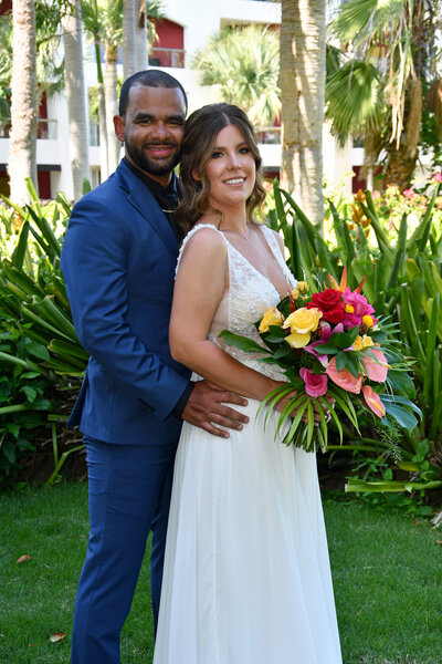 A smiling bride and groom pose in a lush garden setting, holding a vibrant bouquet after their tropical destination wedding.