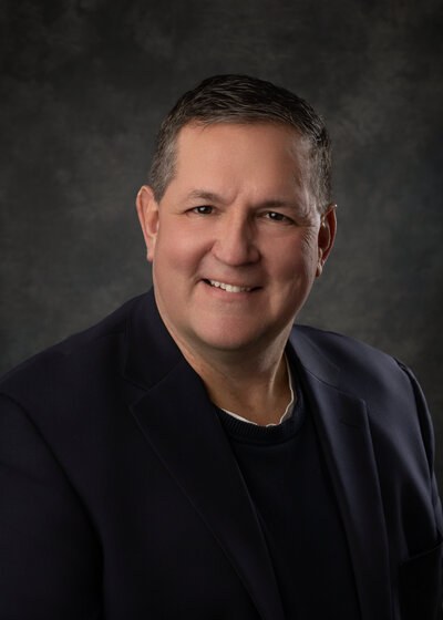 A man wearing black sits in front of a grey background for his Iowa headshot photographer.