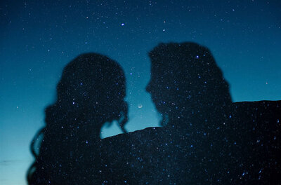 A couple's silhouetted profiles are reflected against a starry night sky, their faces nearly touching. The deep blue hues and scattered stars create a dreamy, celestial effect.