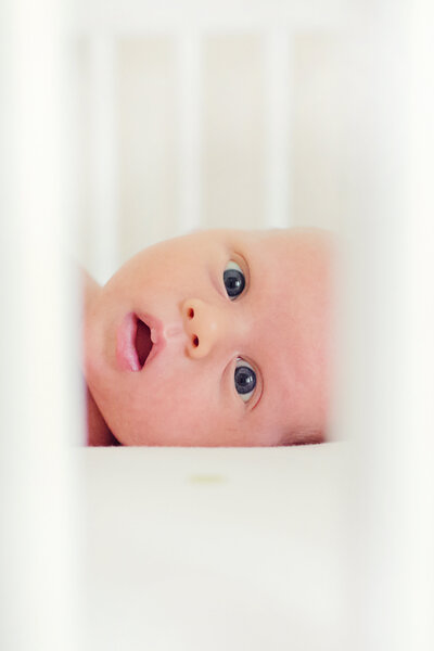 Baby photographed with wide open eyes looking through the slats of the crib.