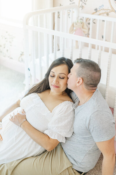 A couple sits in a nursery in front of a white crib. The pregnant wife leans against her husband and closes her eyes. Her husband kisses her on the cheek, captured by a Leander Maternity Photographer.
