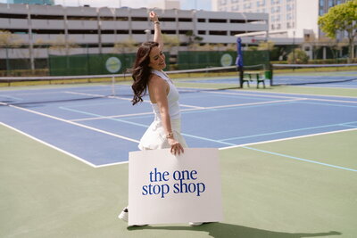 Woman on tennis court holding sign saying the one stop shop