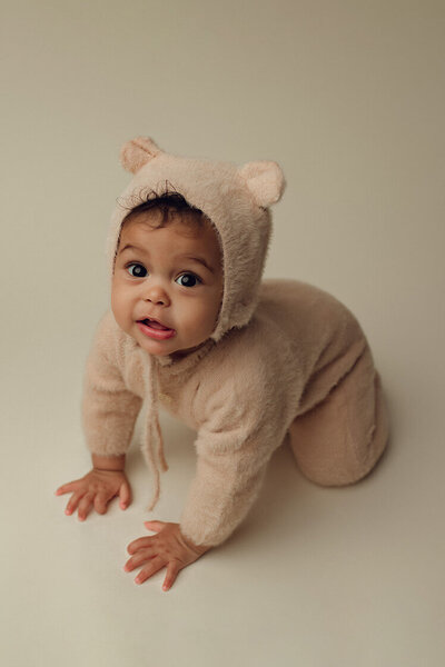 6 month old girl on a cream backdrop wearing a fuzzy cream teddy outfit