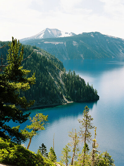 A tranquil lake surrounded by dense, green pine trees, with a mountain featuring patches of snow in the background. The clear blue water reflects the sky, and the scene conveys a sense of calm and pristine natural beauty.