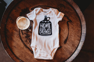 onesie that says "Home Brewed" sitting on a whiskey barrel with a glass of beer beside it