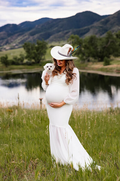 Pregnant woman in a white dress and cowboy hat holds her belly and her small white dog against a background on mountains and a lake.